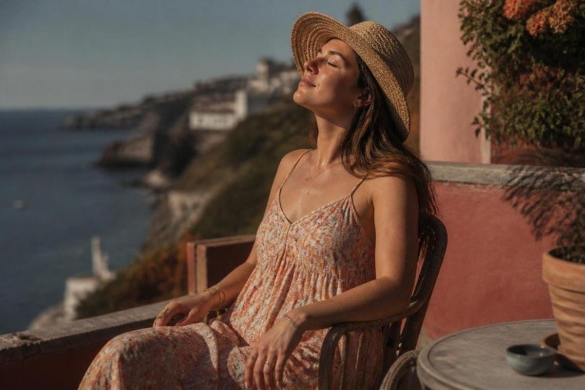 Μια γυναίκα με ψάθινο καπέλο και κλειστά μάτια απολαμβάνει τον ήλιο σε μια βεράντα πάνω από τη θάλασσα.